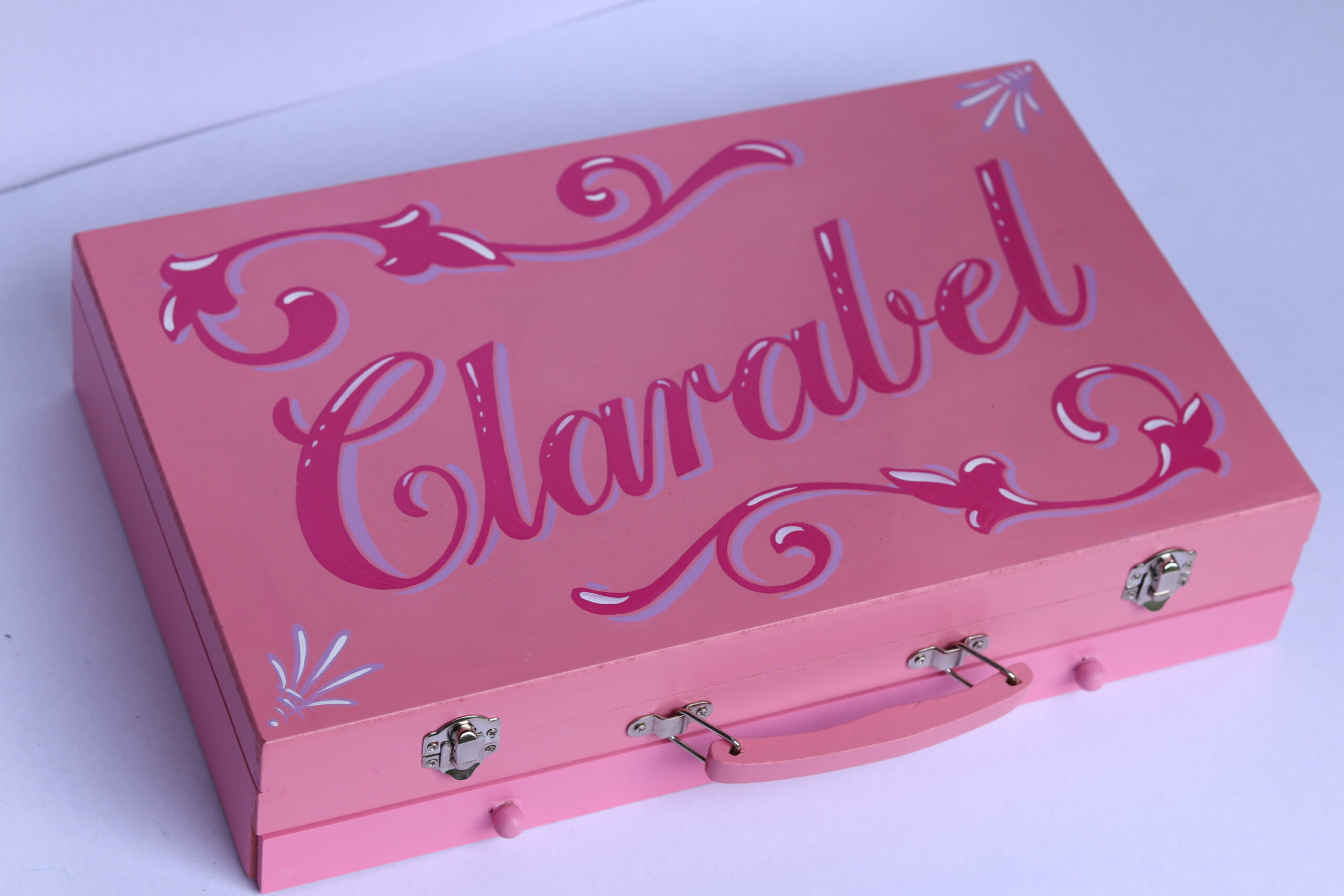 The name Clarabel in hot pink with violet and white details and scrollwork  painted onto the top of a light pink art box 