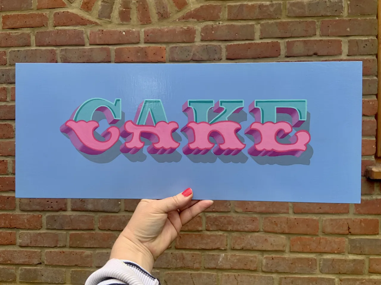 hand holding a handpainted sign with the word CAKE in pinks, blues and violet enamel paints on aluminium panel